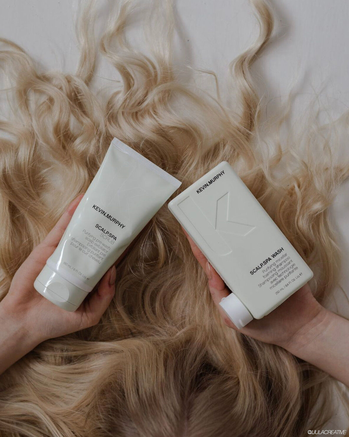 Two bottles of Kevin Murphy scalp care products held over a background of wavy blonde hair.